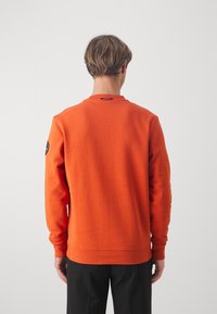Felpa arancione con collo rotondo e polsini a costine. Presenta una piccola patch con logo sulla manica sinistra. Il materiale sembra morbido e testurizzato.
