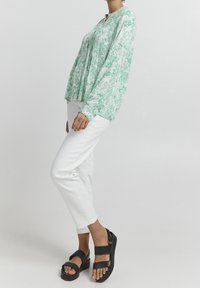 Long-sleeved white blouse with green floral pattern, featuring a pleated front and a round neckline. Worn with white pants and black sandals.