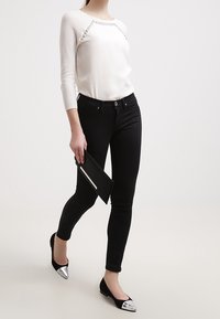 Bílý top s texturovanými rukávy a kovovými akcenty, černé úzké džíny a černé baleríny se stříbrnými špičkami, držící černé psaníčko.