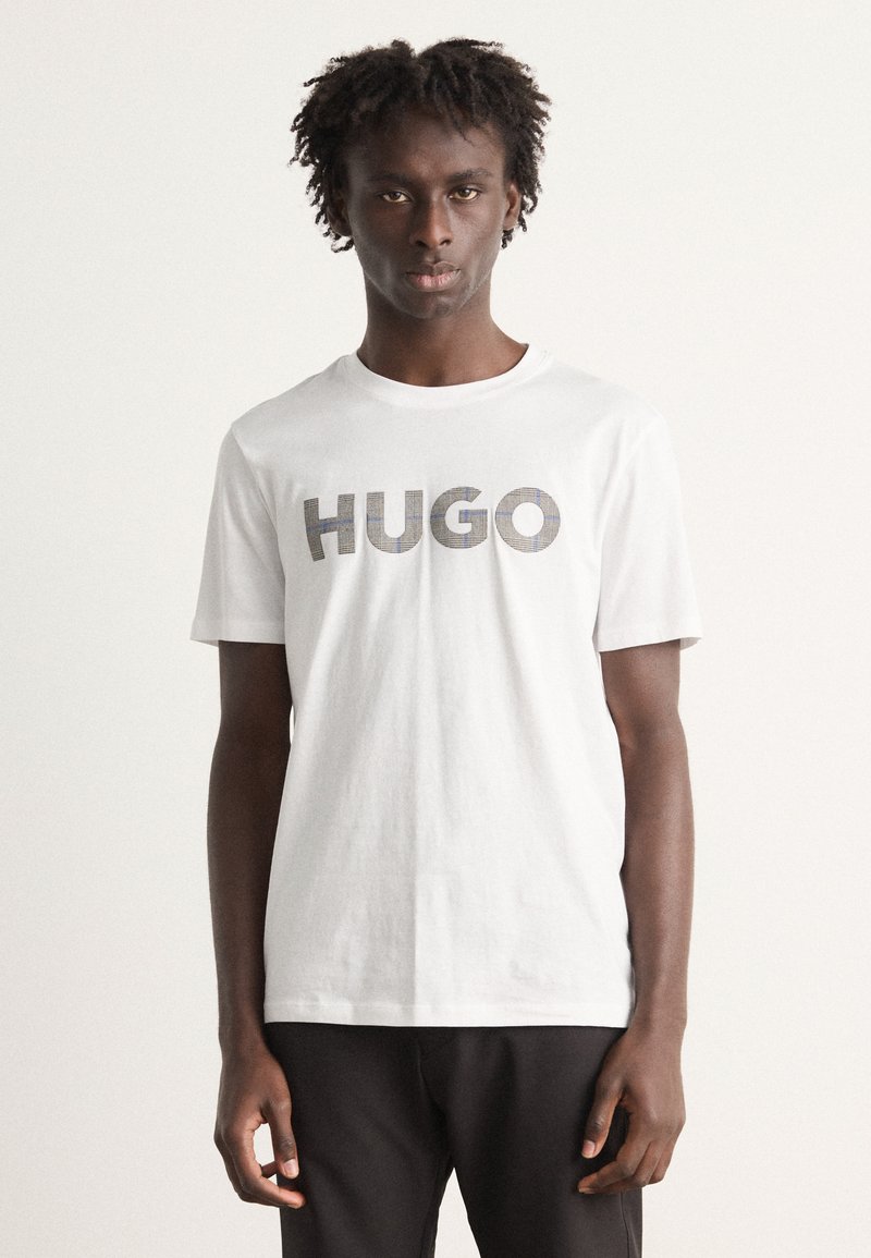Hvit bomulls t-skjorte med rund hals, med et grått mønstrete "HUGO"-logo på forsiden. Korte ermer, avslappet passform.