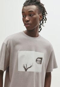 Lys grå bomulds t-shirt med grafisk tryk af et træ og et rundt vindue, med korte ærmer og rund halsudskæring.