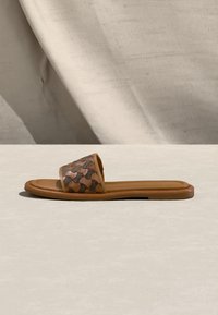 Sandale à glissière marron avec semelle rembourrée et bride en cuir tissé à motifs, posée sur une surface beige clair avec un fond en tissu ombragé.