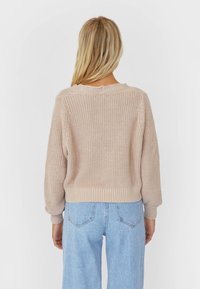 Mujer rubia vestida con un suéter de punto beige y unos jeans de tiro alto azul claro, de pie con la espalda hacia la cámara sobre un fondo blanco.