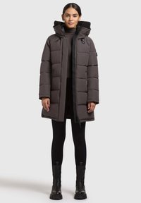 Abrigo acolchado en gris oscuro, con capucha, cierre de cremallera y textura acolchada. Combinado con leggings negros y botas negras gruesas.