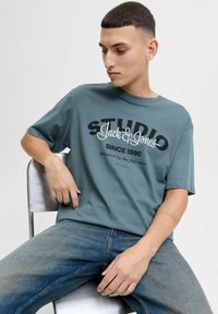 Teal bomulds T-shirt med korte ærmer, der har et stort sort og hvidt grafisk print med teksten "STUDIO Jack & Jones" og noget andet tekst nedenfor.