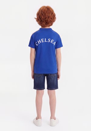 Enfant aux cheveux roux portant un maillot de sport Chelsea bleu, un short en jean foncé et des baskets blanches, debout de dos sur fond blanc.