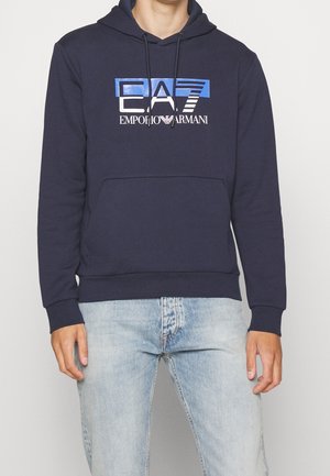 Man wearing navy blue Emporio Armani hooded sweatshirt with front pocket and light blue jeans against a plain background.