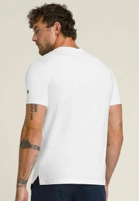 Hombre con cabello rizado y tatuajes en los brazos, vistiendo una camiseta de manga corta blanca y sencilla, de espaldas, contra un fondo claro y liso.