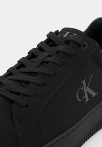 Zwarte sneaker met een gladde leren textuur, ronde neus, veters en een rubberen zool. Heeft een grijs "cK" logo aan de zijkant.