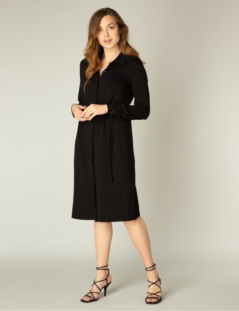 Zwarte button-up jurk met lange mouwen, voorzien van een strikceintuur en midi-lengte. Draagt met zwarte decolleté hakken. Gladde stof en klassiek ontwerp.