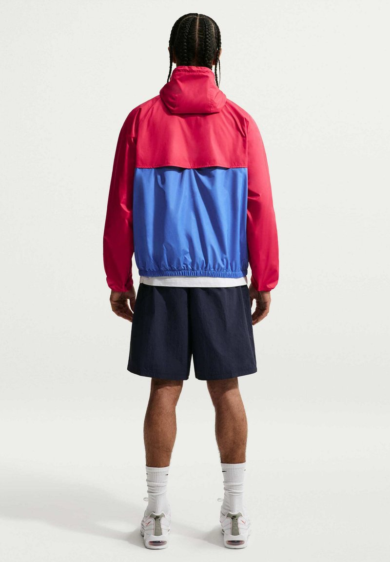 Man met gevlochten haar, gekleed in een rood-blauw capuchonjack, zwarte shorts, witte sokken en witte sneakers, staand met zijn rug naar de kijker.