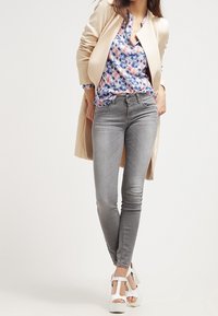 Jasno beżowy długi płaszcz, niebieska i różowa bluzka w wzory, szare obcisłe dżinsy oraz białe platformy. Skupienie na warstwowaniu i nowoczesnym kroju.