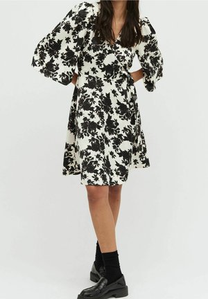 Frau in einem weißem Wickelkleid bis zum Knie mit schwarzem Blumenmuster, Puffärmeln, schwarzen Socken und schwarzen Loafers, stehend mit Händen in die Hüften gestützt.