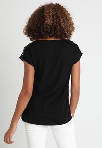 Černé tričko s krátkými rukávy, kulatým výstřihem a volným střihem. Obsahuje dekorativní knoflíkové detaily na rameni a měkkou texturu tkaniny.