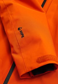 Lys orange jakke med en glat, mat finish. Har et sort logo og velcro-detaljer på ærmerne for sikker lukning.