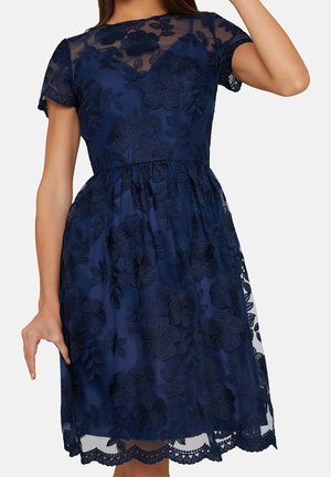 Mujer con un vestido azul marino hasta la rodilla con superposición de encaje floral, mangas cortas transparentes y cintura entallada.