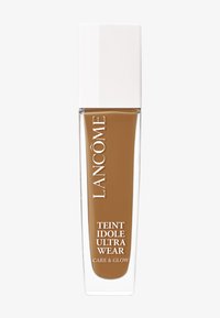Base de maquillaje Lancome Teint Idole Ultra Wear en un frasco de vidrio transparente con una tapa blanca alta. El color es un tono marrón cálido.