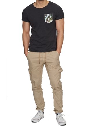 T-shirt à manches courtes gris foncé avec une poche à motif de palmiers, associé à un pantalon cargo beige et des baskets blanches. Coupe décontractée, style casual.