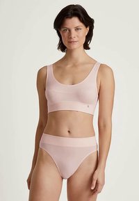 Rövid, sötét hajú nő, aki világos rózsaszín seamless melltartót és hozzá illő magas derekú fehérneműt visel, egy sima háttér előtt áll.