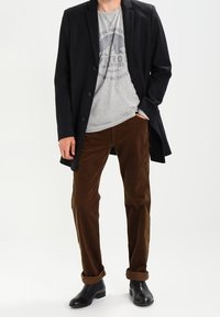 Svart ylleblandad kappa, grå grafisk T-shirt, bruna kordflortorsbyxor och svarta läderstövlar anklar. Avslappnad outfit med varierande texturer.