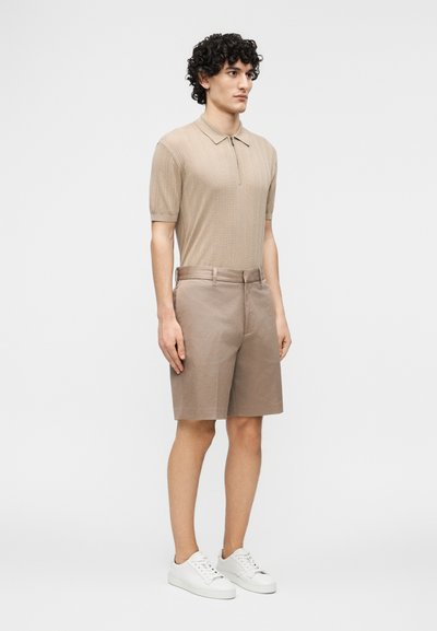Άνδρας με κατσαρά μαλλιά που φοράει μπεζ κοντομάνικο πόλο πουκάμισο, σορτς μέχρι το γόνατο στο ίδιο χρώμα και λευκά αθλητικά παπούτσια, στέκεται μπροστά σε απλό φόντο.