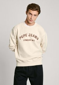 Sudadera beige con la inscripción "PEPE JEANS LONDON W11" en burdeos. Presenta un cuello redondo y un ajuste cómodo y oversized. Combinada con pantalones oscuros.