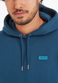 Blue hoodie with a drawstring hood, featuring a small blue TOG24 label. Soft fabric with a smooth texture and metallic aglets on the drawstrings.