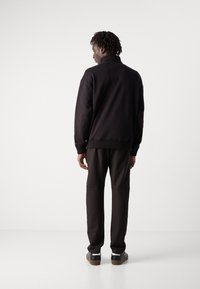 Sudadera negra con cuello y puños acanalados, combinada con pantalones oscuros y zapatos negros, vista desde atrás contra un fondo claro.