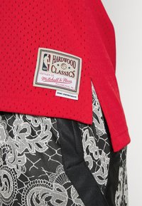 Punainen verkkopaita, jossa on "HARDWOOD CLASSICS" -merkki, yhdistettynä mustiin shortseihin, joissa on valkoinen kukkakuvio ja nauhat.