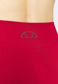 Leggings sportivi rossi con una vita alta texture. Un accento con logo grigio è posizionato vicino alla parte superiore della vita.