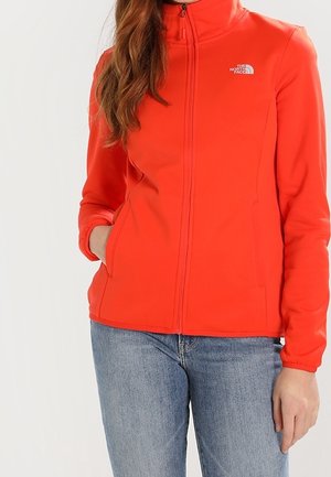 Nainen pukeutunut kirkkaan oranssiin vetoketjulliseen takkiin, jossa on The North Face -logo, yhdistetty sinisiin farkkuihin, seisoo yhdellä kädellä taskussa.