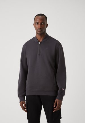 Sweat-shirt gris foncé avec un col zippé à quart, des poignets côtelés et un petit logo sur la poitrine. Matière douce et texturée.