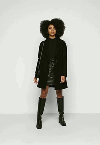Joven mujer con cabello rizado y voluminoso, vestida con abrigo negro, falda de cuero negro, botas hasta la rodilla y llevando un bolso negro con cadena.