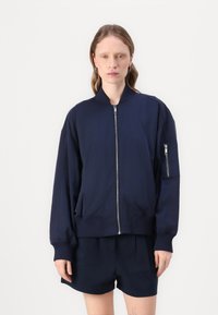 Μπλε ναυτικού bomber jacket με μπροστινό φερμουάρ, ριμπ μανσέτες, πλάγιες τσέπες και τσέπη με φερμουάρ στο μανίκι. Λείο φινίρισμα υφάσματος και χαλαρή εφαρμογή.