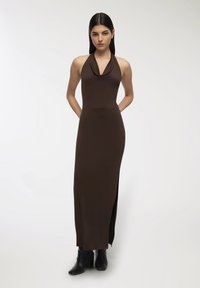 Abito maxi marrone con scollo a cowl, silhouette aderente e alta spaccatura laterale; abbinato a stivaletti neri alla caviglia.