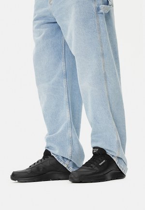 Czarne sportowe trampki z teksturowaną skórzaną cholewką, okrągłym noskiem i wyściełanym kołnierzem; noszone z jasnoniebieskimi, luźnymi dżinsami.