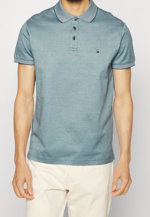 Mann in einem taillierten blauen Polohemd mit gestreiftem Kragen und Ärmelsäumen, kombiniert mit hellbeigen Hosen, steht mit entspannten Armen.