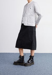 Persona con chaqueta acolchada de color gris claro, falda midi negra con bolsillo y botas de plataforma negras con cordones, de pie sobre una alfombra azul.