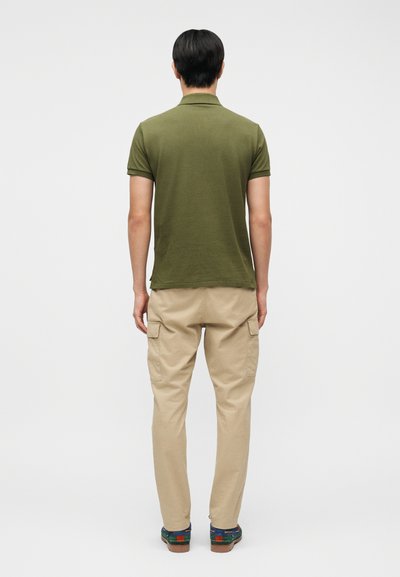 Άνδρας που στέκεται με την πλάτη, φορώντας ένα πόλο πουκάμισο ελιάς, μπεζ παντελόνι καρό και μπλε παπούτσια με καφέ σόλες.