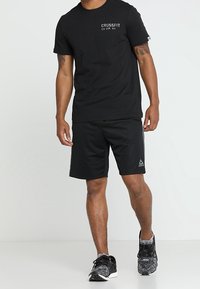 Černé bavlněné tričko s logem "CROSSFIT", spárované s černými sportovními šortkami a vzorovanými teniskami, na neutrálním pozadí.