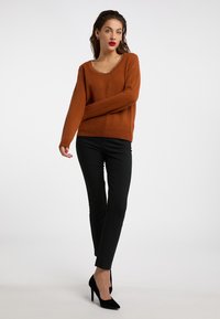Brændt orange sweater med rund halsudskæring og guld kæde detalje, parret med tætsiddende sorte bukser og sorte højhælede sko.