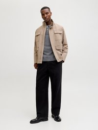 Mann trägt eine beige Jacke über einem grauen Henley-Pullover, schwarze weite Hose und schwarze Brogue-Schuhe, steht vor einem schlichten hellen Hintergrund.