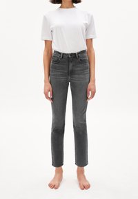 Femme portant un T-shirt blanc uni et un jean noir délavé taille haute, debout pieds nus sur un fond blanc.