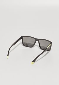 Ochelari de soare dreptunghiulari negri cu lentile gri, având rame din cauciuc. Tempele au vârfuri galbene și accente hardware subtile.