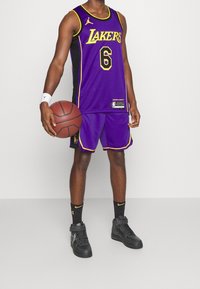 Purppurainen koripallopaita, jossa lukee "LAKERS" keltaisella, mustat yksityiskohdat, yhdistelee shortsit, pitää kädessään koripalloa, parina mustat lenkkarit ja sukat.