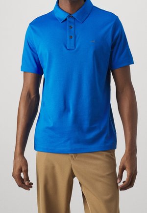 Mann trägt ein leuchtend blaues kurzärmeliges Poloshirt mit drei Knöpfen und hellbraune Hosen, gezeigt vom Hals bis zur Taille.