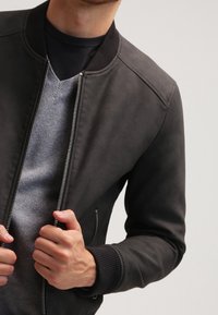Veste bomber noire avec col et poignets côtelés. Fermeture zippée à l'avant, texture lisse, portée sur un pull gris à col en V.