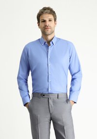 Camicia azzurra chiara a maniche lunghe con colletto a bottone, abbinata a pantaloni grigi. Tessuto morbido, vestibilità strutturata e leggera lucentezza.