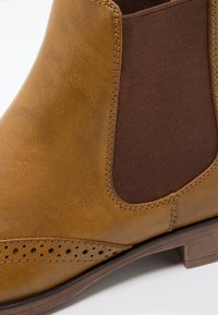 Botte Chelsea en cuir marron avec perforations brogue, panneau latéral élastique, détails de couture et talon bas empilé sur fond blanc.