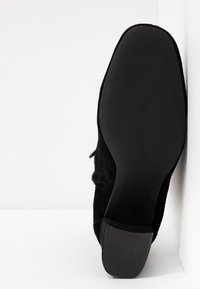 Bottine en suède noir vue de la semelle latérale avec un talon bas et carré sur fond blanc.